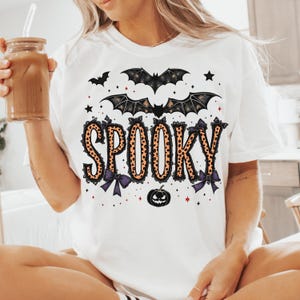 May include: White t-shirt with the word "SPOOKY" in orange leopard print letters, adorned with black bows and bats. A black pumpkin and stars complete the Halloween design. The shirt is a casual, festive garment.