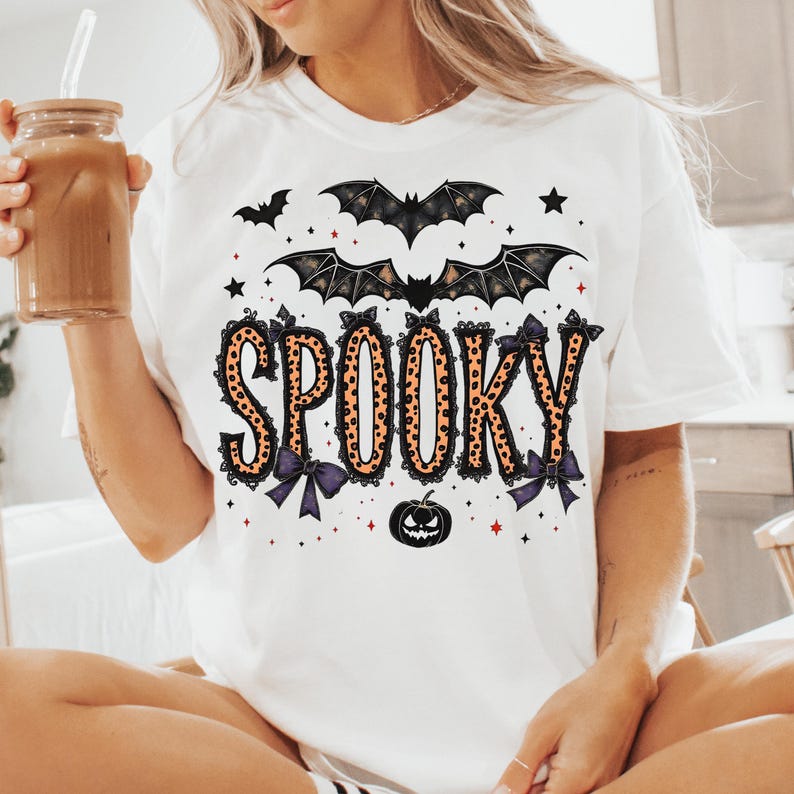 May include: White t-shirt with the word "SPOOKY" in orange leopard print letters, adorned with black bows and bats. A black pumpkin and stars complete the Halloween design. The shirt is a casual, festive garment.
