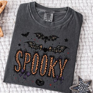 May include: Dark gray t-shirt with a Halloween-themed design. The word "SPOOKY" is printed in orange leopard print with black lace trim and purple bows. Bats, stars, and a pumpkin are also featured in the design.