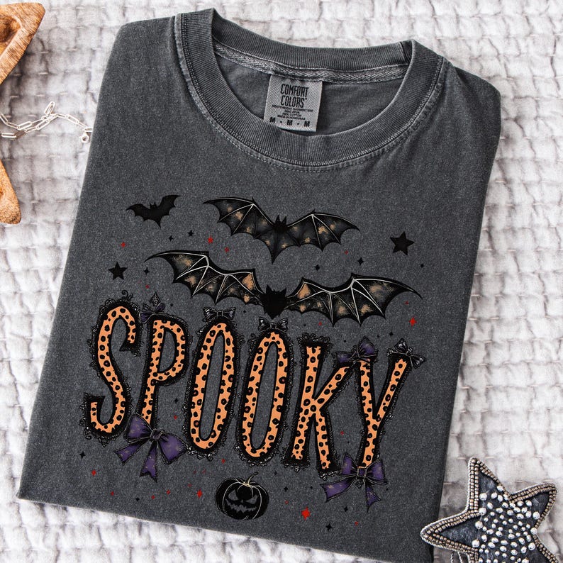 May include: Dark gray t-shirt with a Halloween-themed design. The word "SPOOKY" is printed in orange leopard print with black lace trim and purple bows. Bats, stars, and a pumpkin are also featured in the design.