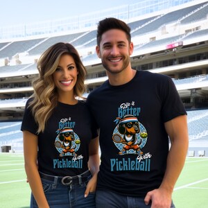 K&ouml;nnte beinhalten: Zwei Personen tragen schwarze T-Shirts mit einem Cartoon-Dackel, der Pickleball spielt. Die Shirts sagen "Life is Better with Pickleball".