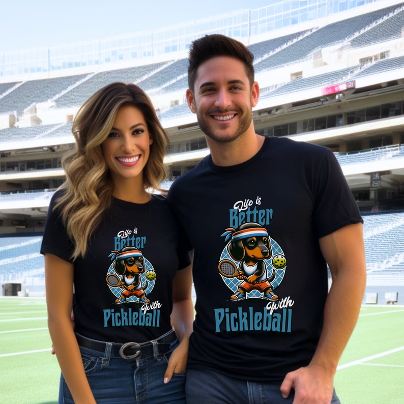 K&ouml;nnte beinhalten: Zwei Personen tragen schwarze T-Shirts mit einem Cartoon-Dackel, der Pickleball spielt. Die Shirts sagen "Life is Better with Pickleball".