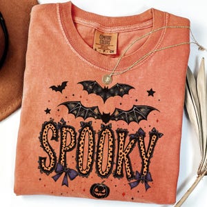 May include: Orange Comfort Colors t-shirt with a Halloween theme. The word "SPOOKY" is printed in a leopard print font with purple bows. Bats, stars, and a jack-o'-lantern are also featured in the design. A gold necklace is draped across the shirt.
