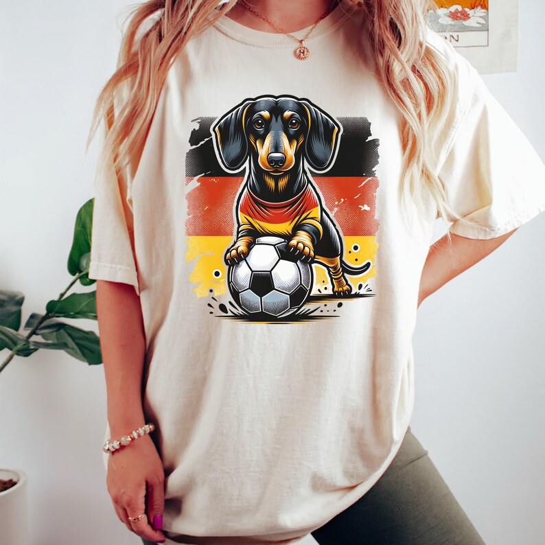 May include: A white t-shirt with a cartoon image of a dachshund wearing a jersey with the German flag colors and holding a soccer ball.