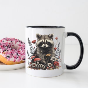 Puede incluir: Taza de cer&aacute;mica blanca con asa y borde negros. La taza presenta una ilustraci&oacute;n detallada de un mapache rodeado de flores y setas. El mapache tiene pelaje gris y negro, y las flores son rojas, amarillas y blancas.