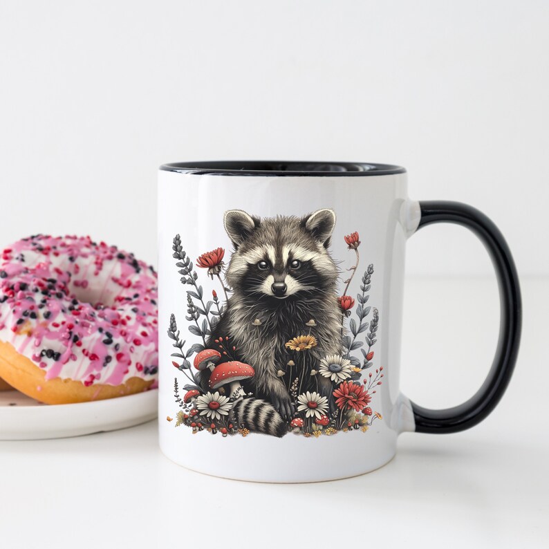 Puede incluir: Taza de cer&aacute;mica blanca con asa y borde negros. La taza presenta una ilustraci&oacute;n detallada de un mapache rodeado de flores y setas. El mapache tiene pelaje gris y negro, y las flores son rojas, amarillas y blancas.