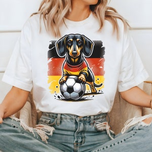 May include: A white t-shirt with a cartoon dachshund wearing a German flag jersey and holding a soccer ball. The background is a distressed German flag.