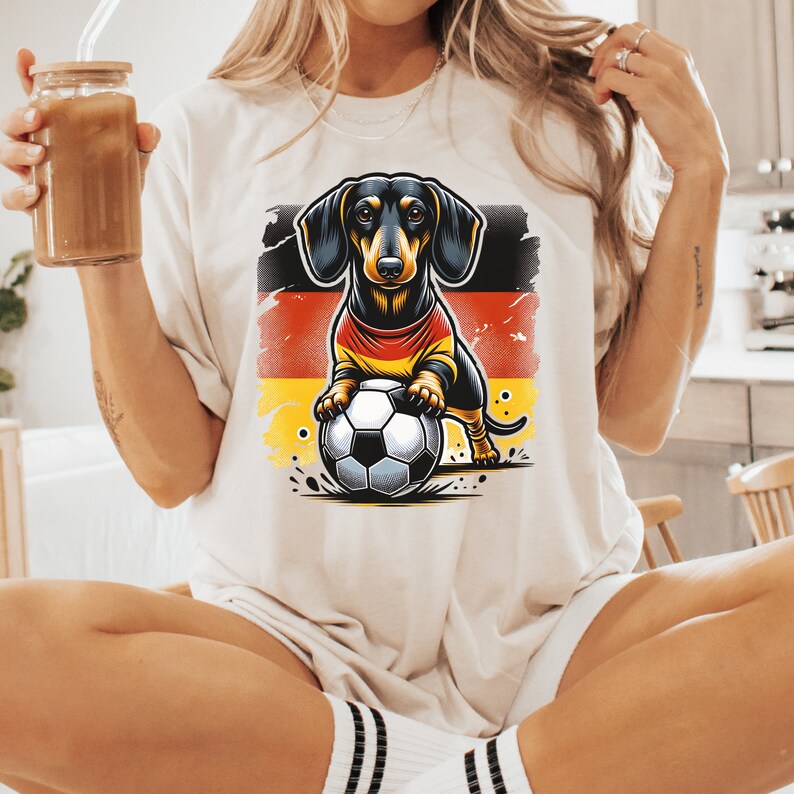 May include: A white t-shirt with a cartoon image of a dachshund wearing a jersey with the German flag colors and holding a soccer ball.