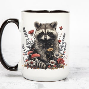Puede incluir: Taza de cer&aacute;mica blanca con asa y borde negros. La taza presenta una ilustraci&oacute;n detallada de un mapache rodeado de flores y setas en tonos rojos, amarillos y blancos. El mapache tiene un patr&oacute;n de pelaje negro y gris.