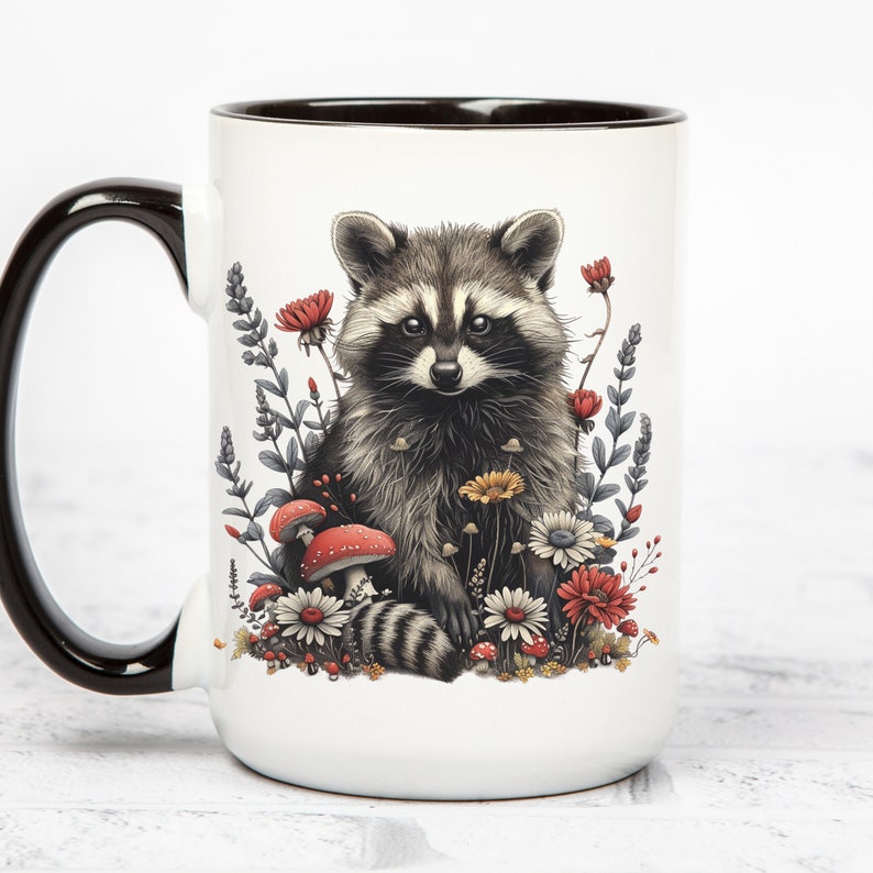 Puede incluir: Taza de cer&aacute;mica blanca con asa y borde negros. La taza presenta una ilustraci&oacute;n detallada de un mapache rodeado de flores y setas en tonos rojos, amarillos y blancos. El mapache tiene un patr&oacute;n de pelaje negro y gris.