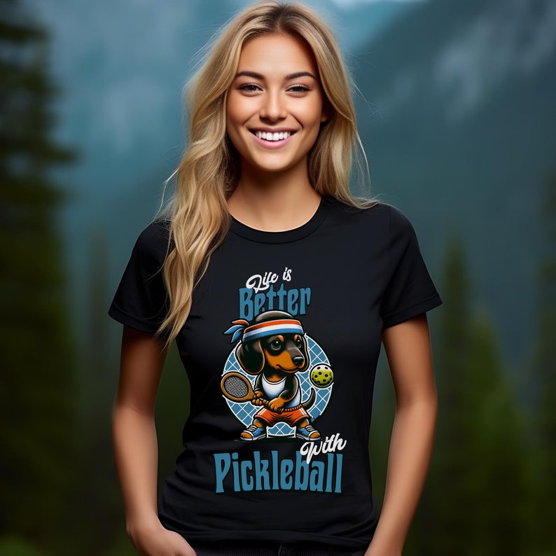 K&ouml;nnte beinhalten: Schwarzes T-Shirt mit einem Cartoon-Dackel, der ein Stirnband tr&auml;gt und einen Pickleball-Schl&auml;ger h&auml;lt. Der Text auf dem Shirt lautet "Life is Better with Pickleball".