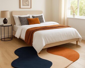 May include: A modern bedroom scene featuring a bed with white bedding, a rust-colored throw, and decorative pillows. A unique, abstract rug in navy blue, beige, and orange sits on the wooden floor. A framed artwork and a side table with a lamp complete the decor.