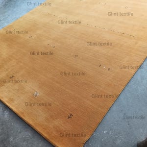 May include: A close-up of a light brown rug with a subtle pattern of small white dots and a few black lines. The rug is made of a soft, textured material and has a slightly faded appearance. The text "Glint textile" is repeated throughout the image.