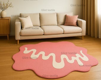 May include: A pink, irregularly shaped rug with a white, wavy design on a hardwood floor. A beige sofa with two pillows and a wooden cabinet are in the background. The room has a neutral color palette.