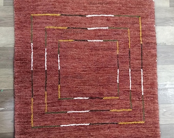 May include: A red rug with a geometric pattern of white, yellow, and brown lines. The rug has a square shape and a textured surface. The text "Glint textile" is visible in the top right corner of the rug.