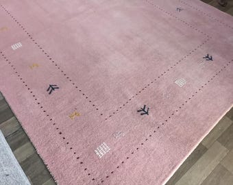 Puede incluir: Una alfombra rosa rectangular grande con un borde de pequeños puntos oscuros y formas geométricas decorativas. La alfombra presenta patrones repetidos en forma de flecha y cuadrados en azul oscuro, blanco y amarillo.