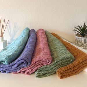 Könnte beinhalten: Fünf gestrickte Decken in Pastellfarben: Hellblau, Lila, Rosa, Grün und Orange. Jede Decke ist gefaltet und zeigt ein strukturiertes, gewebtes Muster. Die Decken sind auf einer weißen Oberfläche angeordnet, mit einer kleinen Pflanze und einem Diffusor im Hintergrund.