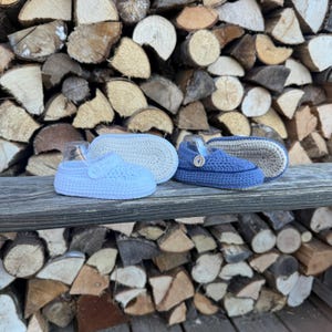 Könnte beinhalten: Drei Paar handgefertigte Häkelschuhe in Hellblau, Off-White und Marineblau. Jeder Schuh hat einen Knopfverschluss und ein strukturiertes Design. Die Schuhe werden auf einer Holzoberfläche vor einem Hintergrund aus gestapeltem Brennholz präsentiert.