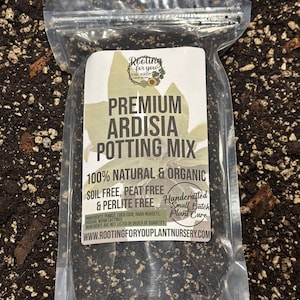 May include: A clear plastic bag filled with a dark brown potting mix labeled "Premium Ardisia Potting Mix". The label also states "100% Natural & Organic", "Soil Free, Peat Free & Perlite Free", and "Handcrafted Small Batch Plant Care". The website address "www.rootingforyouplantnursery.com" is printed on the label.