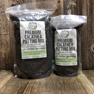 May include: Two bags of potting mix labeled "Premium Calathea Potting Mix" with the text "Soil Free & Peat Free" and "100% Natural and Organic". The bags are on a wooden surface.