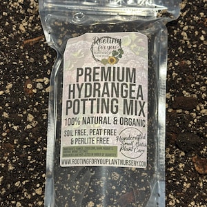 May include: A clear plastic bag filled with a dark brown potting mix for hydrangeas. The bag has a white label with the text "Rooting for you Plant Nursery Eugene Oregon", "Premium Hydrangea Potting Mix", "100% Natural & Organic", "Soil Free, Peat Free & Perlite Free", "Handcrafted Small Batch Plant Care", and "www.rootingforyouplantnursery.com".