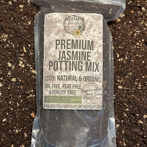 May include: A clear plastic bag filled with dark potting mix. The label reads "Premium Jasmine Potting Mix" and includes text about being 100% natural and organic, soil-free, peat-free, and perlite-free. The bag is on a dark brown surface.