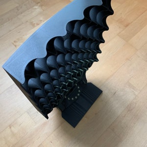 Puede incluir: Un objeto negro impreso en 3D con un diseño ondulado. El objeto está formado por muchas piezas pequeñas que se superponen y crean una superficie texturizada.
