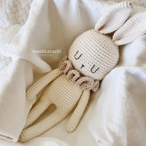 Może przedstawiać: Pluszowa zabawka króliczka wykonana na szydełku z długimi uszami, falbanką i haftowanymi oczami. Króliczek leży w białym koszyku.