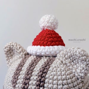 tabby crochet amigurumi santa cat sitting. Cat in red santa hat entering the chimney. 100% cotton yarn beige brown tabby cat Christmas crochet softie on the white christmas tree background.