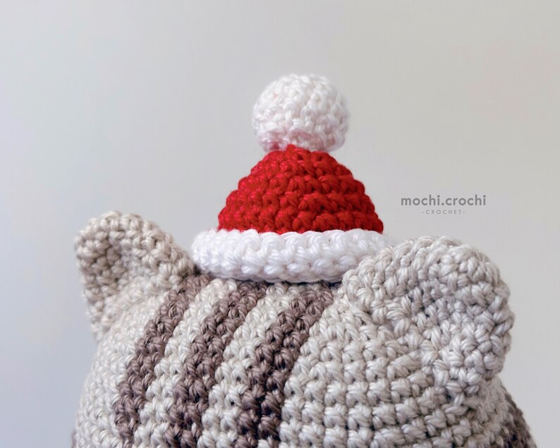 tabby crochet amigurumi santa cat sitting. Cat in red santa hat entering the chimney. 100% cotton yarn beige brown tabby cat Christmas crochet softie on the white christmas tree background.