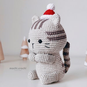 tabby crochet amigurumi cat sitting. Cat in red santa hat entering the chimney. 100% cotton yarn beige brown tabby cat crochet softie on the white christmas trees background.