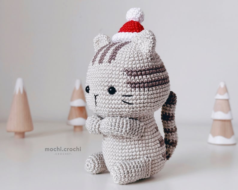 tabby crochet amigurumi cat sitting. Cat in red santa hat entering the chimney. 100% cotton yarn beige brown tabby cat crochet softie on the white christmas trees background.