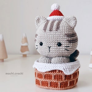 tabby crochet amigurumi santa cat sitting in the chimney. Cat in red santa hat entering the chimney. 100% cotton yarn beige brown tabby cat Christmas crochet softie on the white christmas trees background.