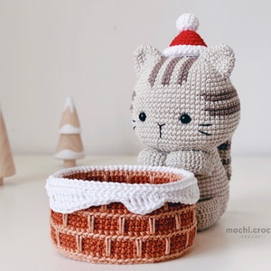 tabby crochet amigurumi santa cat sitting behind the chimney. Cat in red santa hat entering the chimney. 100% cotton yarn beige brown tabby cat Christmas crochet softie on the white christmas tree background.