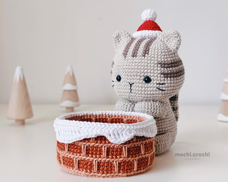 tabby crochet amigurumi santa cat sitting behind the chimney. Cat in red santa hat entering the chimney. 100% cotton yarn beige brown tabby cat Christmas crochet softie on the white christmas tree background.