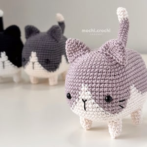 May include: Three crocheted cat plushies in different colors: black, gray, and purple. The purple cat is in focus and has a white face with black eyes and whiskers. The text "mochi.crochi CROCHET" is visible in the background.
