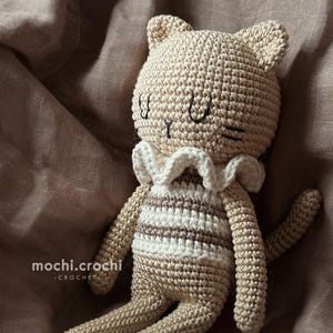 Può includere: Un giocattolo di peluche a forma di gatto con una maglietta a righe bianche e marroni e un colletto bianco all'uncinetto. Il gatto è sdraiato su uno sfondo di tessuto marrone.