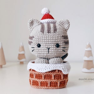 tabby crochet amigurumi santa cat sitting in the chimney. Cat in red santa hat entering the chimney. 100% cotton yarn beige brown tabby cat Christmas crochet softie on the white christmas trees background.