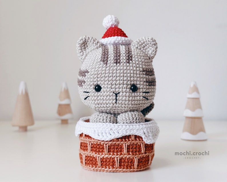 tabby crochet amigurumi santa cat sitting in the chimney. Cat in red santa hat entering the chimney. 100% cotton yarn beige brown tabby cat Christmas crochet softie on the white christmas trees background.