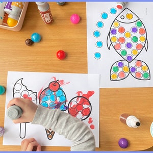May include: A child is using dot markers to color in a picture of an ice cream cone and a popsicle. The picture is on a white sheet of paper. There are also dot markers and a fish picture on the table.