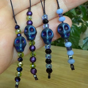 May include: Four beaded skull charms in shades of blue, purple, and green. Each charm features a metallic blue skull bead, accented with various colored beads. The charms are strung on black cords, creating a unique and spooky accessory.