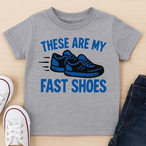 May include: A gray short-sleeved t-shirt with the text "THESE ARE MY FAST SHOES" in blue. The shirt features a graphic of two blue and black sneakers with motion lines. White sneakers and blue jeans are also visible.