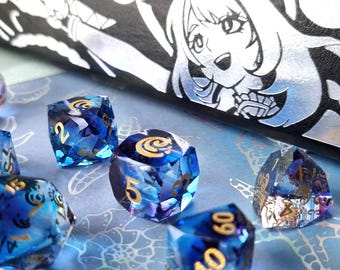 May include: A collection of blue and gold polyhedral dice with a fantasy theme. The dice are set against a blue background with a decorative box featuring a silver anime-style illustration. The dice have gold numbering.