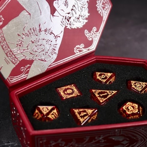 May include: A red coffin-shaped box with a silver illustration on the lid, containing seven red and gold dice. The dice are arranged in a black foam insert. The box is open, displaying the dice.