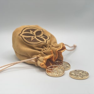 Puede incluir: Una bolsa de gamuza marrón con un diseño bordado en marrón y un cierre de cordón. La bolsa está abierta y se derraman monedas de oro con un diseño similar.