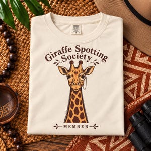 May include: A cream-coloured t-shirt featuring a brown graphic of a giraffe wearing a monocle. The text above the giraffe reads Giraffe Spotting Society and the text below reads MEMBER. The shirt has a crew neck and a Comfort Colors brand label.