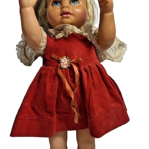 May include: Vintage doll with blonde hair and blue eyes, wearing a red velvet dress with a bow and floral embellishment. The doll has its arms raised, and is wearing a lace collar and sleeves. A classic toy.