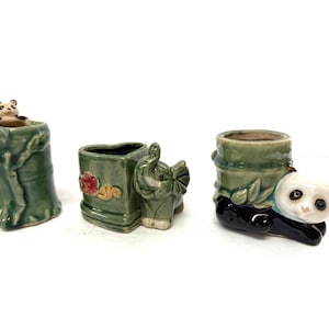 May include: Three small, green ceramic planters. One is shaped like a tree trunk with a panda peeking out. Another is a square planter with an elephant. The third is a bamboo-style planter with a panda.
