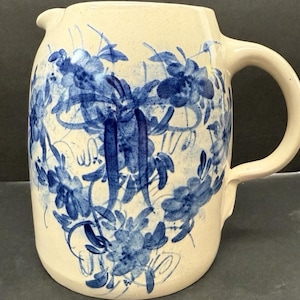 May include: Cream-colored ceramic pitcher with a blue floral design. The pitcher has a curved handle and a spout for pouring. The blue floral pattern covers the body of the pitcher, creating a vintage aesthetic.