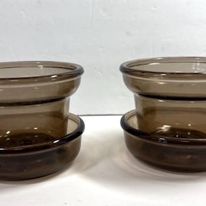 May include: Two matching smoke-colored glass vessels, each featuring a wide, flared upper rim and a separate, rounded base component. The glass is translucent and dark brown in tone, with a smooth, polished finish on both pieces.